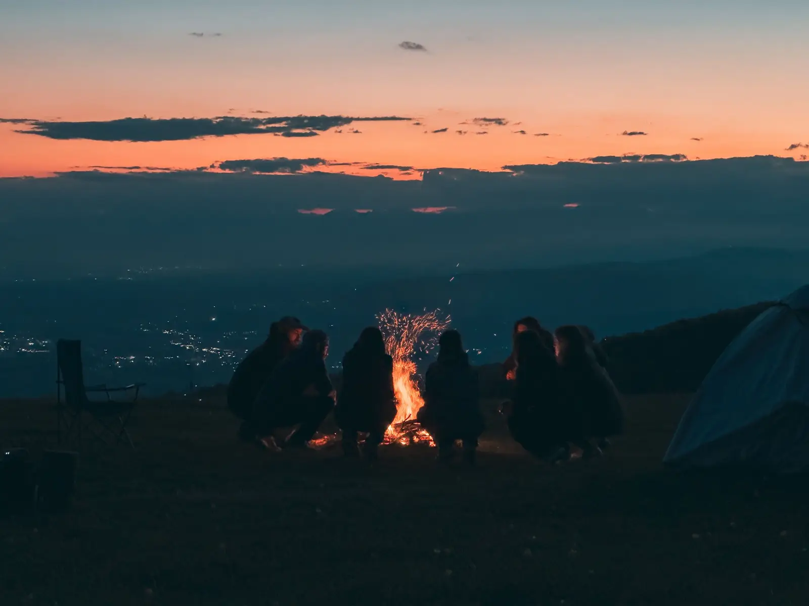 Group of friends crouched around a campfire on a hillside at dusk, valley lights below